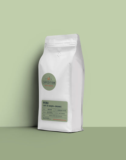 A white stand-up coffee bag labeled Expedition Peru Café de Mujer-Organic, standing on a matching light green surface against a pale green background.